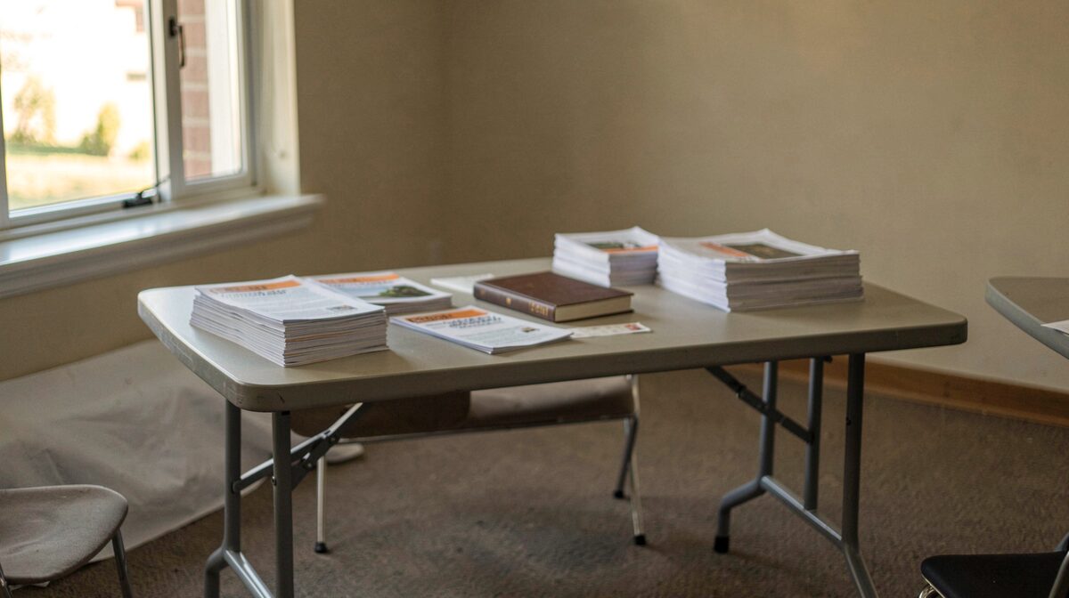 A plain folding table at the back of a meeting hall, late afternoon light through a window, with pamphlets stacked in rows and a closed book beside them; straight-on framing.